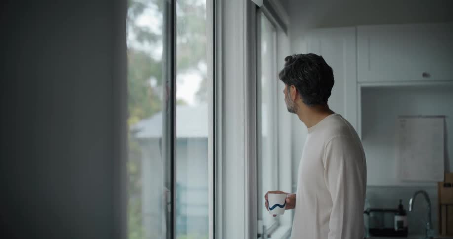 A man holding a cup looks out the kitchen window at home