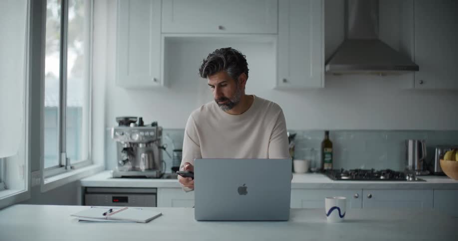 A man sits at his kitchen counter and looks at his phone while on his laptop