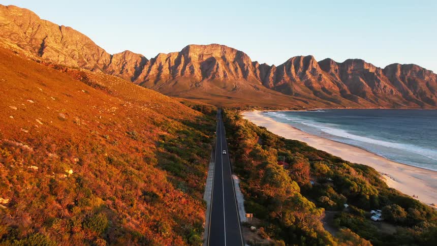 A large asphalt road with a car passing through a mountainous region with a beach next to it