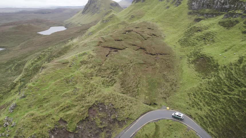 An aerial view reveals a winding road snaking its way through a dramatic landscape of rolling hills and rocky cliffs with a car making its way along the scenic route