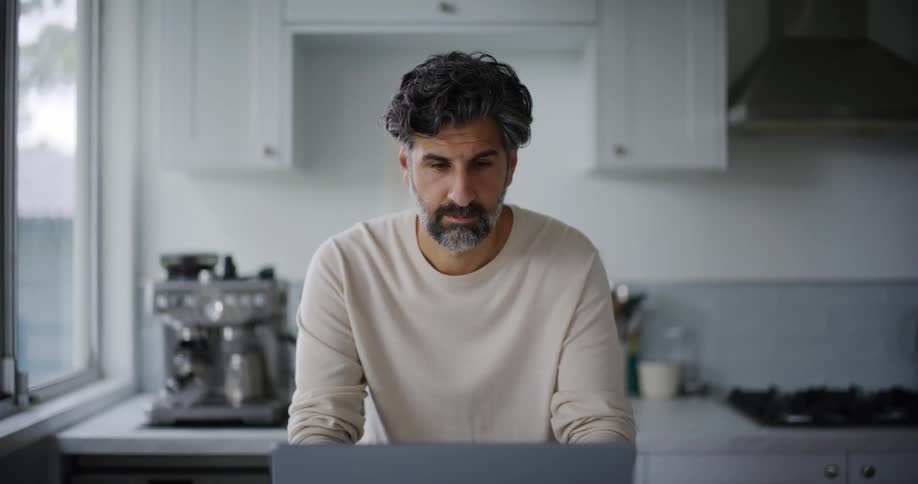 A bearded man wearing a cream sweatshirt smiles at the camera while using a laptop in the kitchen
