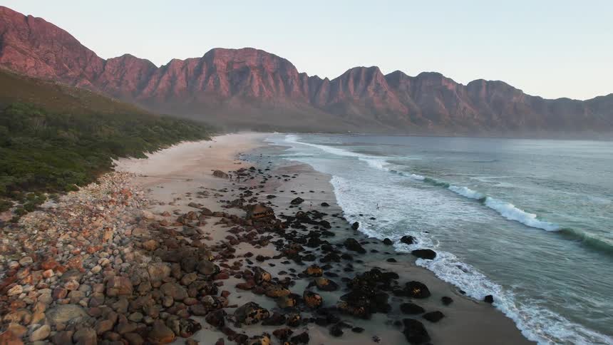 A rocky beach with waves crashing against it stretches along the coastline with dramatic mountains in the distance and a few people enjoying the scenic view