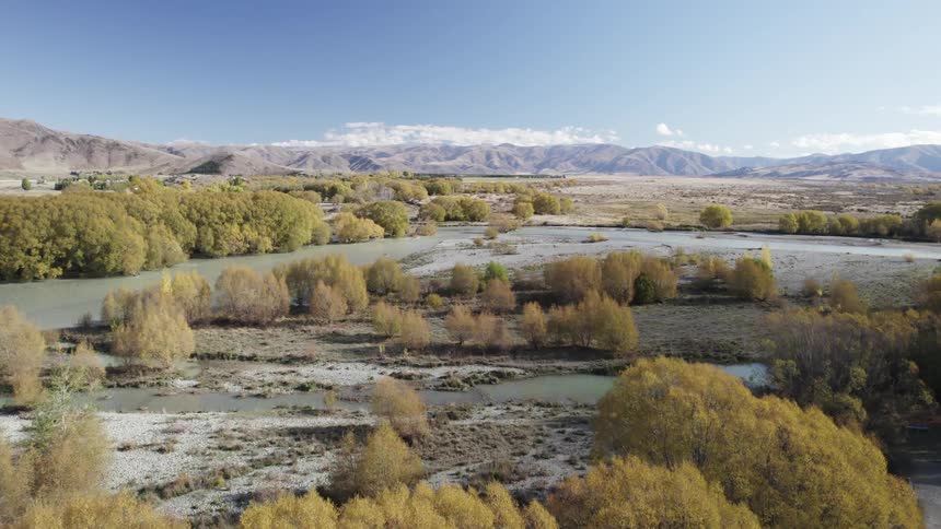 A river meanders through a valley its banks lined with vibrant yellow autumn foliage while distant mountains rise in the background under a clear blue sky