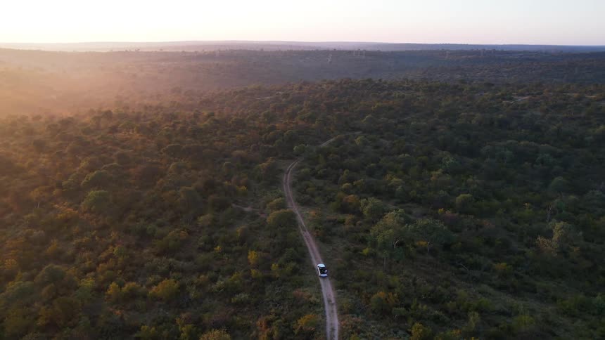 A white vehicle driving along a dirt road through a vast densely forested area at sunrise