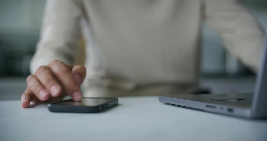 A man working on a laptop reaches out to scroll on the screen of a smartphone on the table