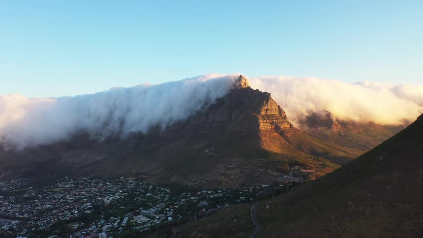 The iconic flattopped peak of table mountain is partially covered by dramatic clouds at sunset casting a shadow over the city below while the remaining peak is bathed in golden sunlight