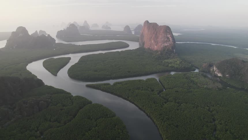 A winding river meanders through a lush green landscape surrounded by dramatic limestone cliffs shrouded in mist