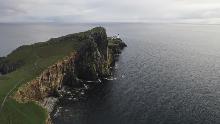 A lighthouse stands on a dramatic cliff overlooking a vast dark ocean on a cloudy day