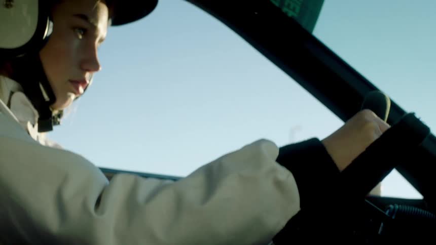 A young woman wearing a helmet and a white jacket is driving a car with one hand on the steering wheel and and changing gears