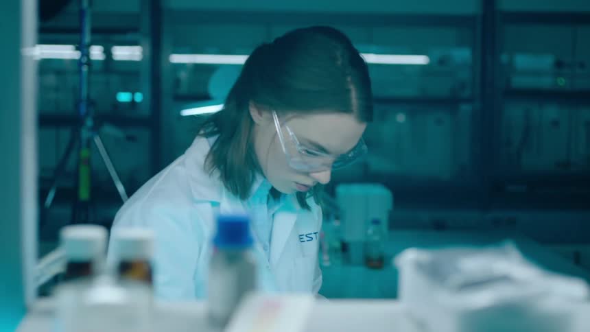 Woman works on a task in a laboratory while a coworker makes notes in the background
