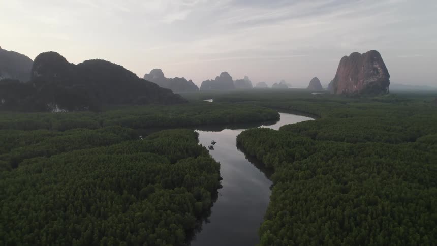 A winding river flows through a dense mangrove forest with dramatic limestone cliffs rising in the background under a hazy sky