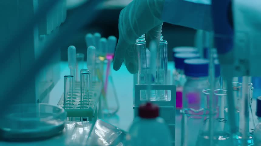 A lab technician places a test tube on a stand in a cluttered workbench
