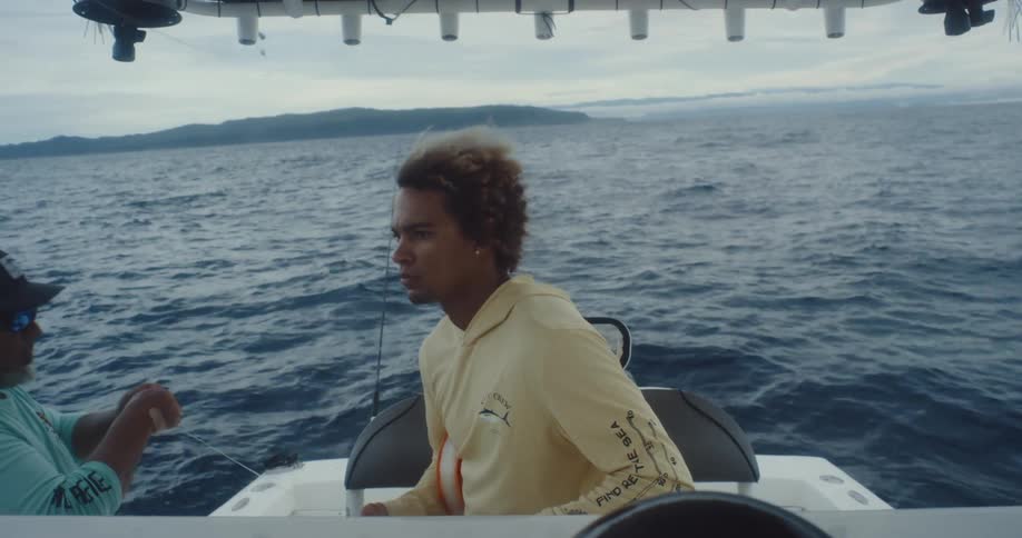 Two people are on a boat in the ocean with one person in a yellow hoodie holding a fishing line while the other sits nearby and a scenic horizon in the background