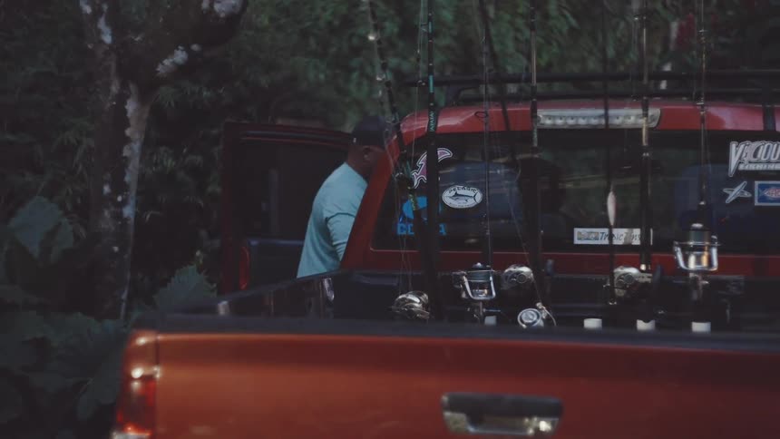 A person in a blue shirt is standing next to a red pickup truck filled with fishing rods and reels