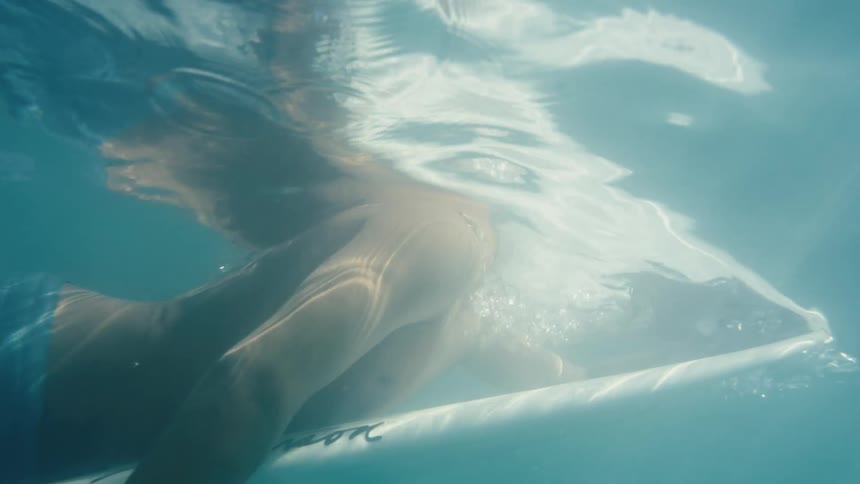 A person is paddling underwater on a surfboard in a clear body of water with bubbles coming from their face