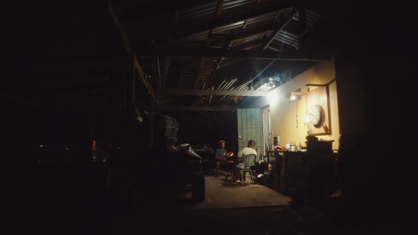 Two men sitting in the covered outdoor area of a house at night with one of them winding a spool of thread