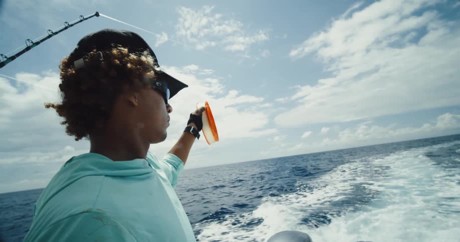 A man wearing sunglasses and a black cap is holding an orange and white object and looking at the waves formed by a boat passing by in an ocean