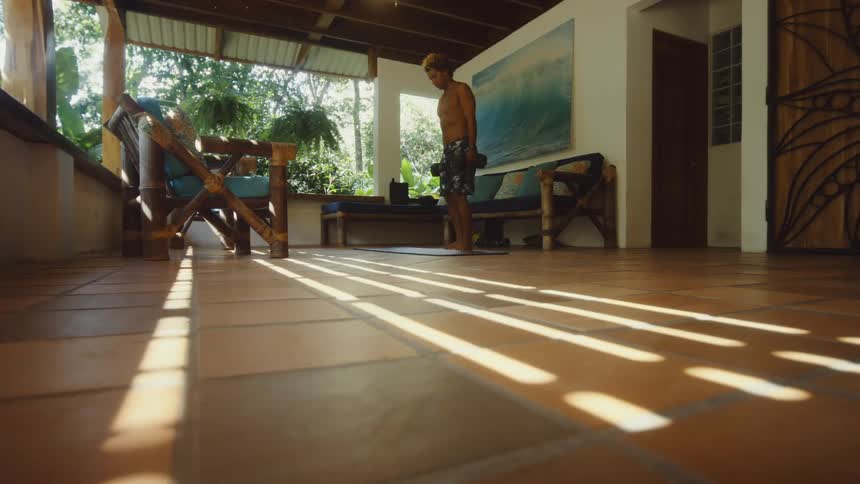 A shirtless man using dumbbells in both hands and he is doing squats on the porch of a house