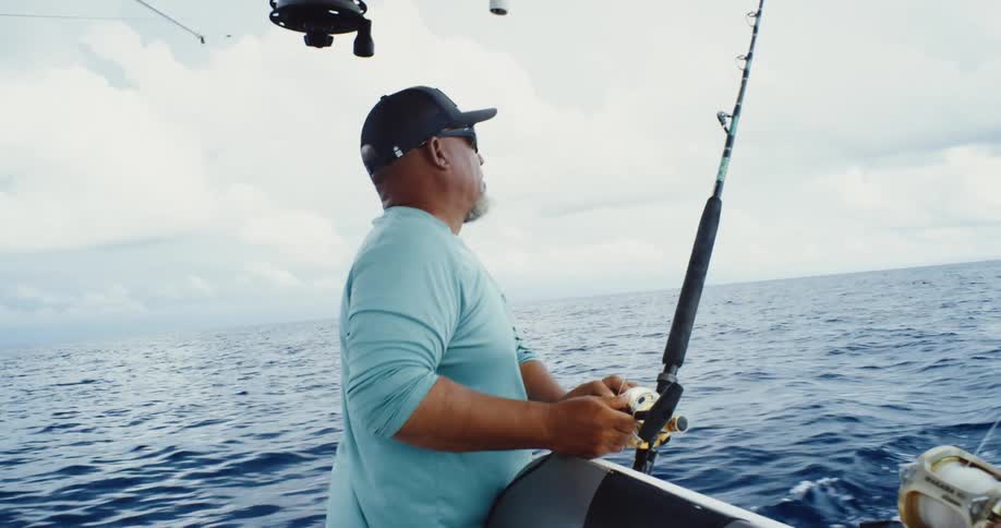 A person is fishing on a boat in the open ocean holding a fishing rod while wearing a light blue longsleeve shirt and a black cap