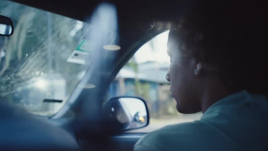 A person with curly hair is shown from the back sitting in the passenger seat of a car looking out the side window as they drive past homes and foliage