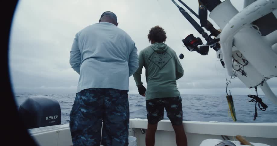 Two men are in a white boat in the ocean and one of them is fishing while the other is handling a fish that is on top of a white box
