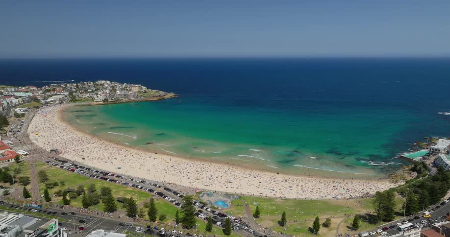 An aerial view of a beach with greenish ocean water and lots of people on the sand and a street crowded with cars next to it