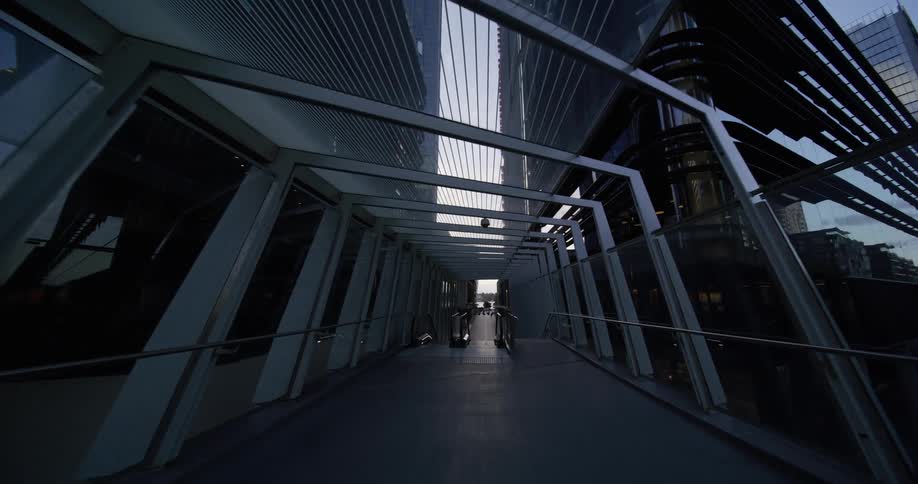 A modern covered walkway with a geometric metal frame leading to a brightly lit exit between tall city buildings