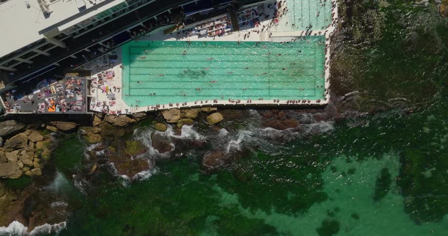 An aerial view of a rectangular pool at a cliffside hotel overlooking the ocean crowded with people bathing