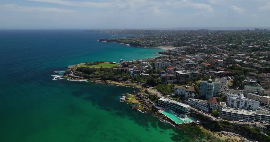Aerial view of the vibrant contrast of turquoise ocean waves crashing against a rocky shoreline juxtaposed with a network of roads and buildings interspersed with lush green parks creating a dynamic coastal cityscape