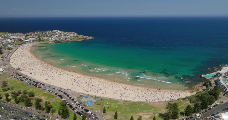 A vast sun drenched beach curves along the coastline dotted with tiny figures enjoying the warm sand and sparkling turquoise water