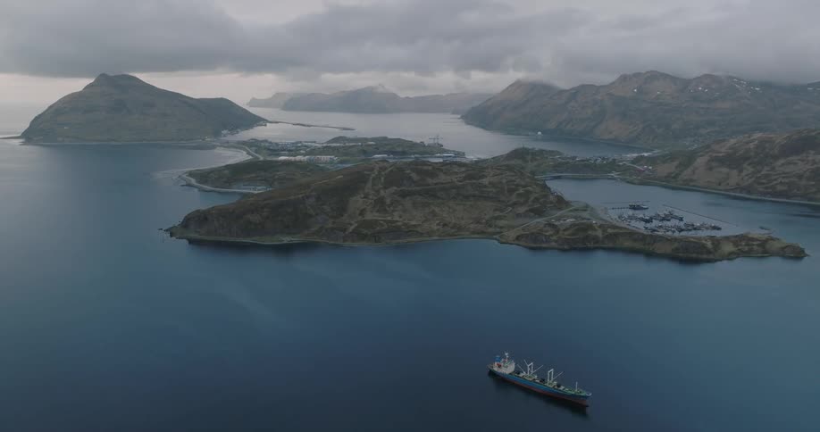 A cargo ship sails through Alaskan bay surrounded by rugged mountains and cloudy skies