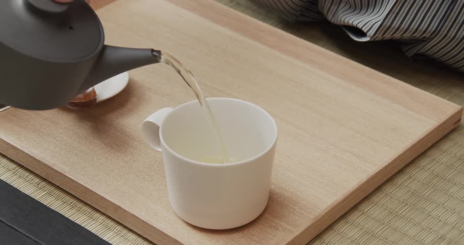 A hand pours tea from a small teapot into a white mug