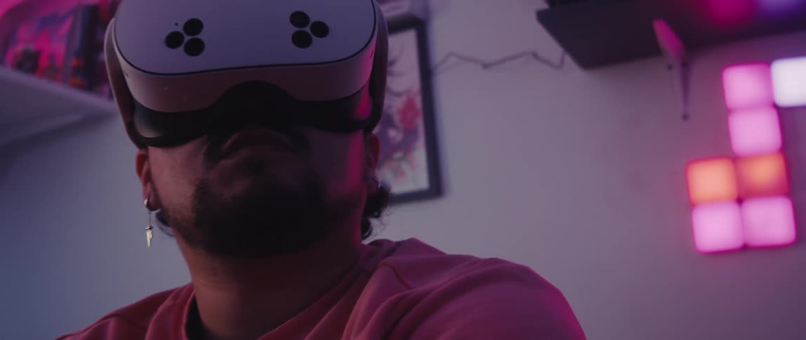A man wearing a white virtual reality headset and a pink t shirt bathed in a purple glow with anime posters on the wall behind him