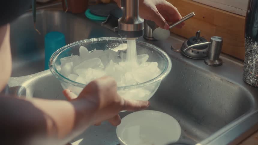 A person is filling a bowl of ice with water in the kitchen sink
