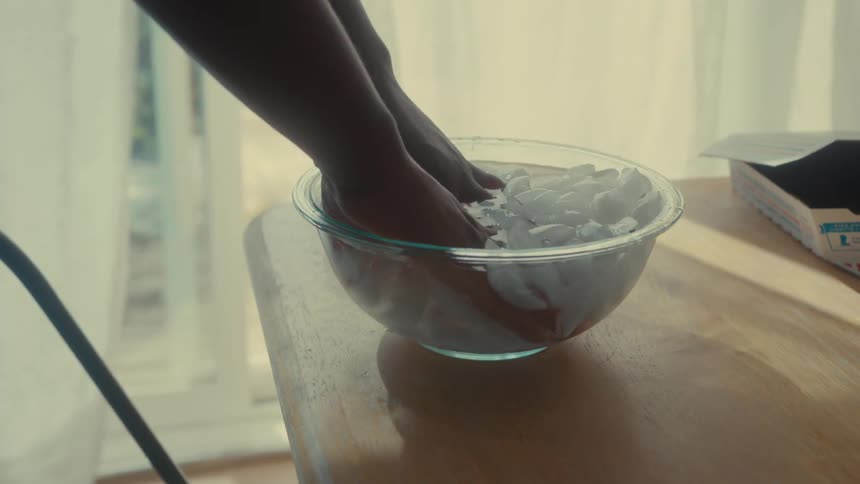 Two hands are submerged in a glass bowl filled with ice cubes on a wooden table
