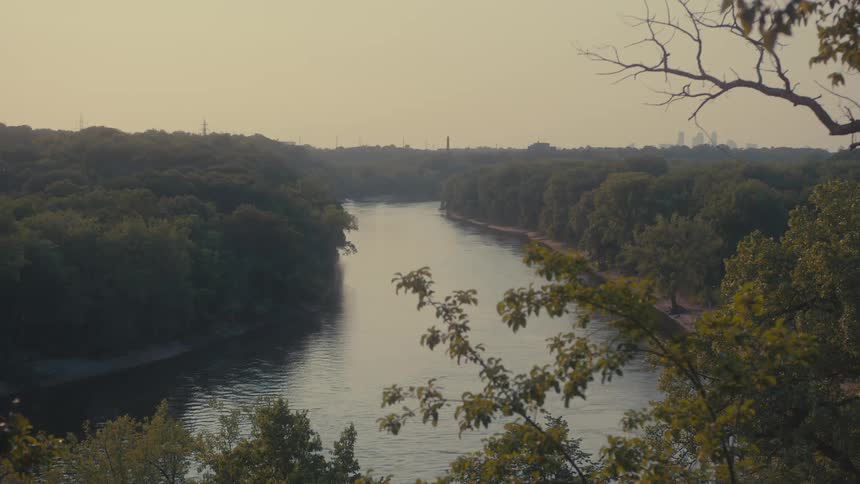 A wide river winds its way through a densely wooded valley with a distant cityscape visible on the horizon