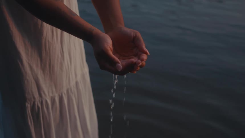 A person in a white dress is holding water in cupped hands with water dripping back into a calm lake