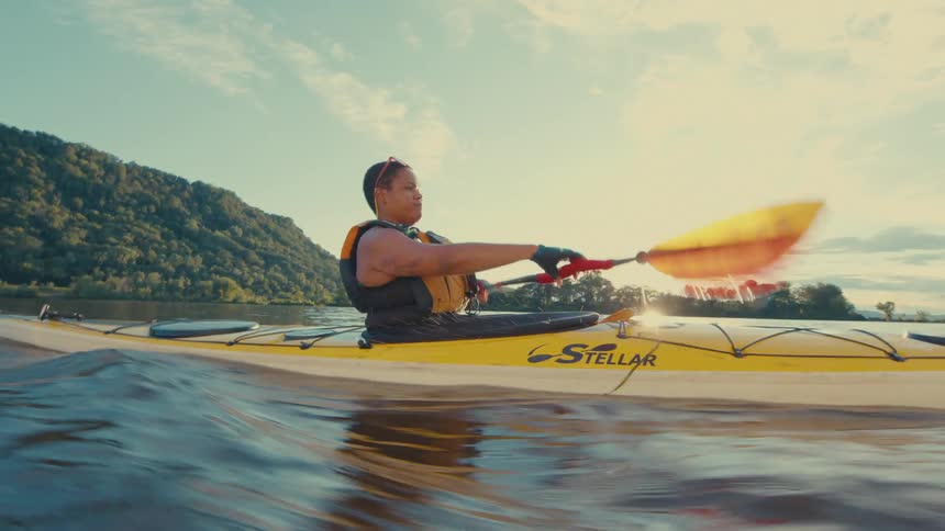 A person wearing a life jacket and gloves is shown kayaking with a red paddle on a body of water with a tree covered shore in the background