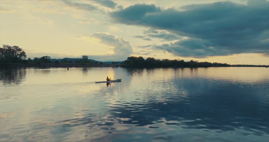 The sky is ablaze with the fiery hues of sunset reflecting in the still water as a kayaker paddles gently their silhouette a dark contrast against the vibrant colors