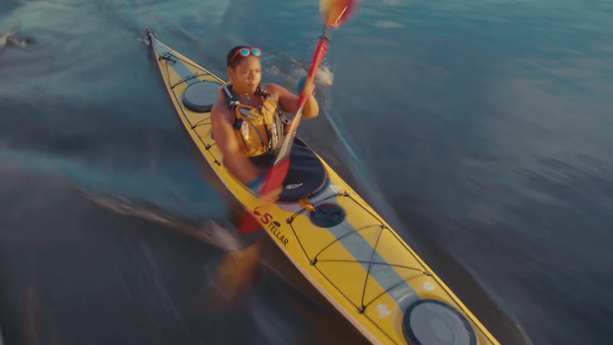 A woman is kayaking on a calm body of water using a doublebladed paddle to propel a yellow kayak forward