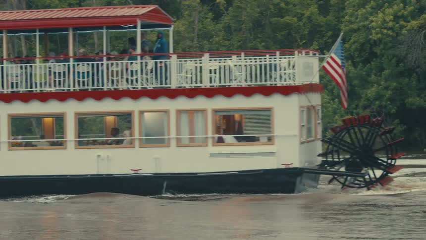 Passengers ride on a riverboat with a paddle wheel past thick woods during the day