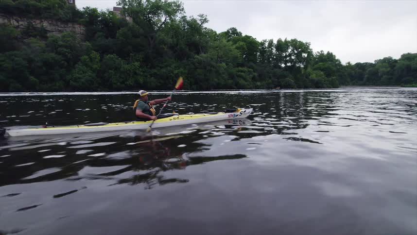 Man kayaks along a river passing through an urban environment on a cloudy day