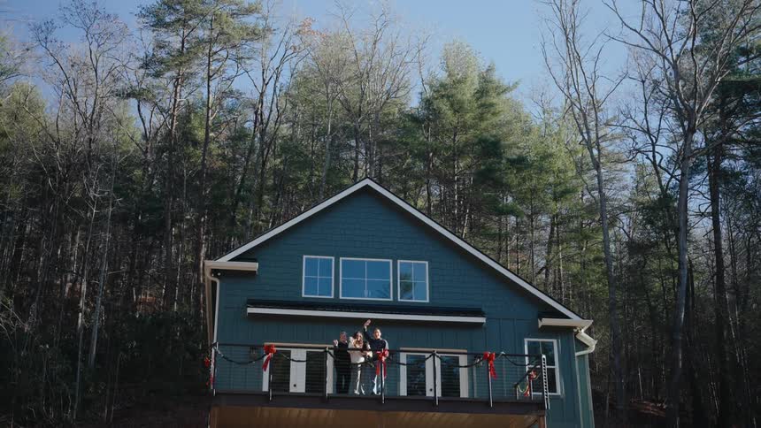 Three people on the balcony of a house located in an environment surrounded by trees making a goodbye sign with their hands