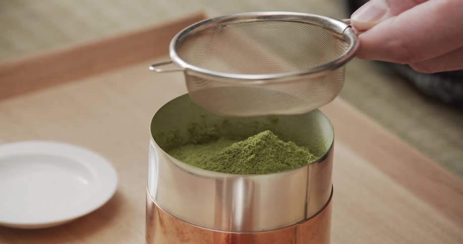 An adult sifts green matcha powdered tea into a metal can