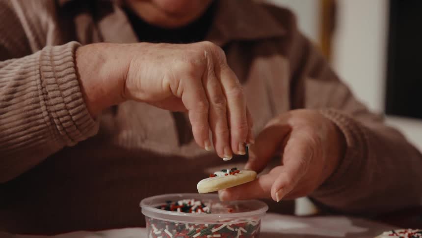Woman decorates sugar cookies with sprinkles while sitting at a table