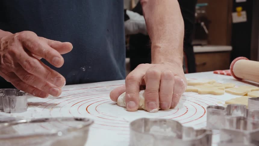 A person is pressing dough with their hand on a floured surface surrounded by various metal cookie cutters and a rolling pin