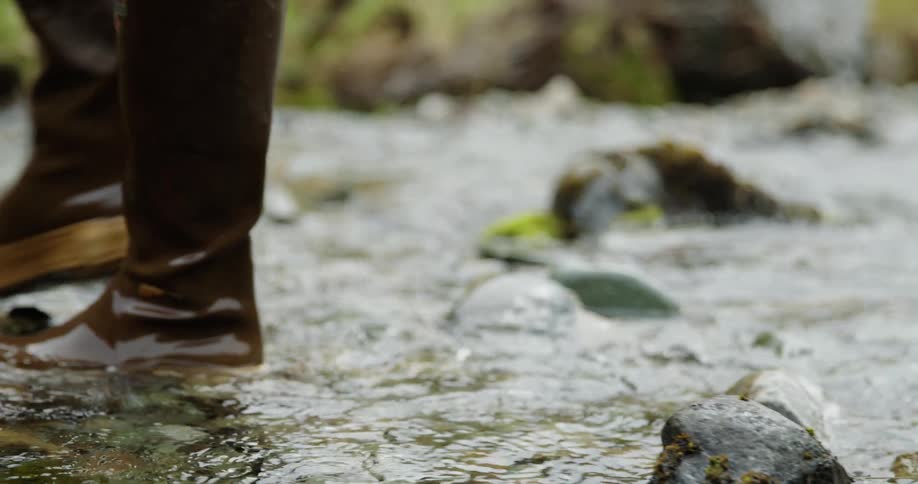 A pair of brown rubber boots walk through a shallow stream splashing water with each step
