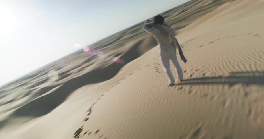 A person dressed in white stands on a sand dune in a vast desert with footprints trailing behind