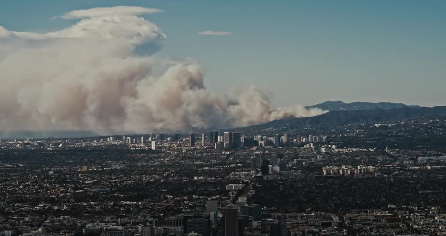 A dense plume of smoke billows over the los angeles skyline obscuring the sun and casting a dark shadow over the city as a wildfire rages in the distant hills