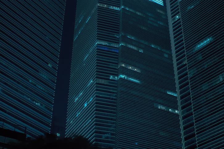 A cluster of towering skyscrapers illuminated with a cool blue light creating a striking contrast against the dark night sky before the lights turn off